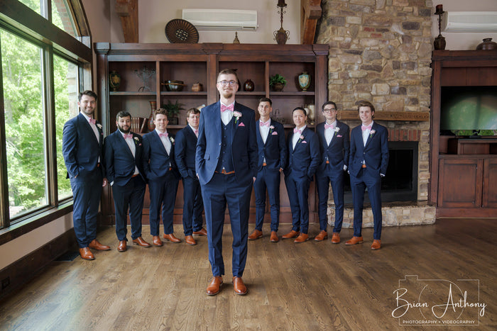 Groom in a navy suit standing in front of his groomsmen, all in navy with pink bow ties